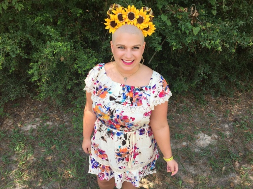 Nicole wearing sunflower ears in a white and flowery dress standing in front of green trees with a large smile on her face as she just lost her fair from chemotherapy.