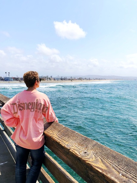 Nicole looking out at the ocean off of a pier with her back facing the camera in a pink Disneyland shirt